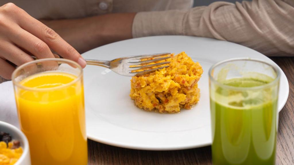 Huevos revueltos con jugo verde y naranja en mesa de desayuno