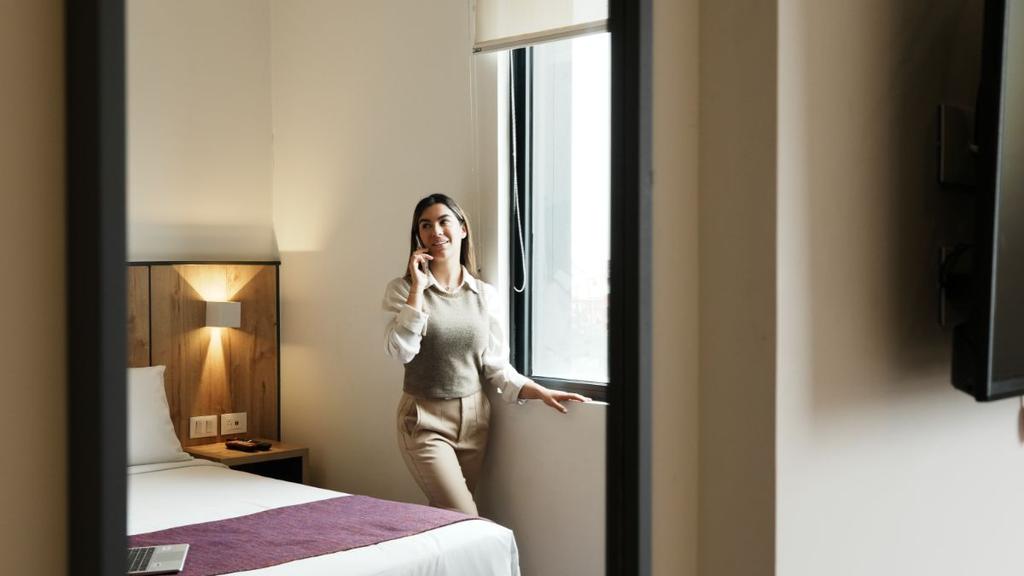 Persona junto a ventana en habitación con cama blanca y mesa de noche
