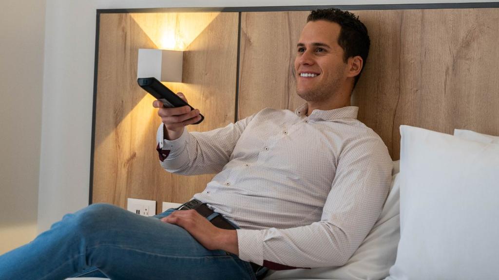 Persona en cama con control remoto y luz cálida sobre cabecera de madera