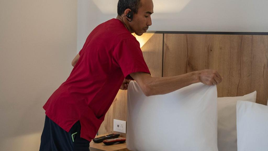 Persona arreglando almohada sobre cama con sábanas blancas y cabecera de madera